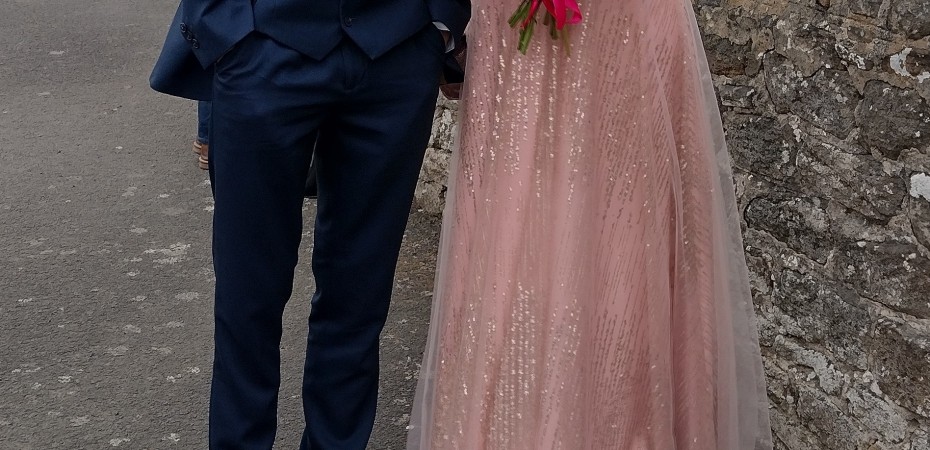 Prom picture of couple. Person on left in navy blue suit. Person on right in long rose gold dress carrying bunch of pink flowers.